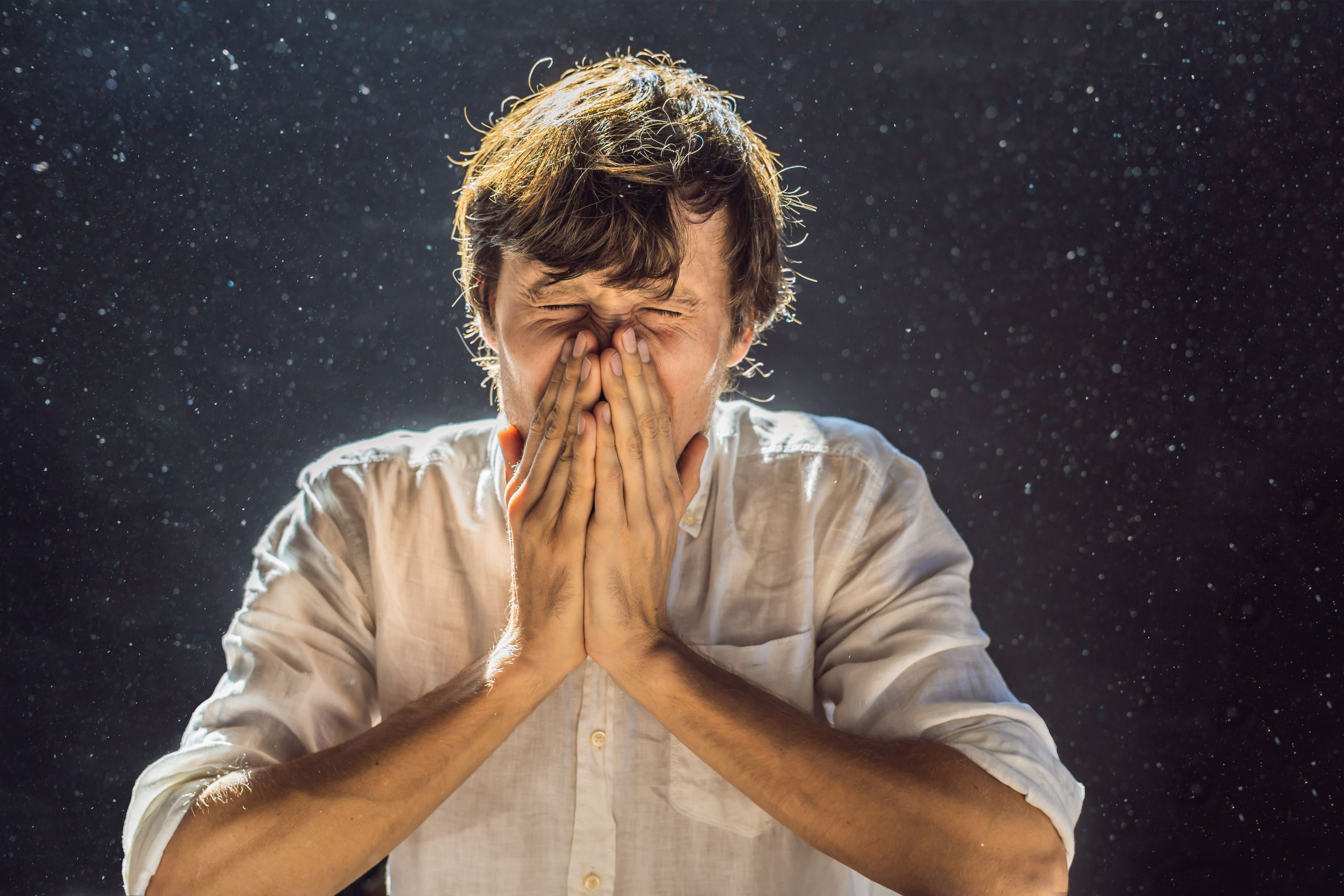 Allergy to dust. A man sneezes because he is allergic to dust. Dust flies in the air backlit by light